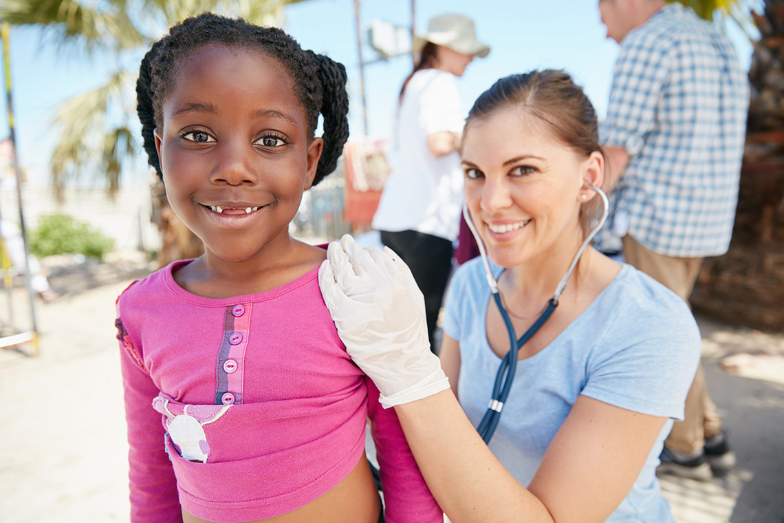 Nurse kneeling down with a stethoscope to listen to the back of a young girl wearing a pink shirt